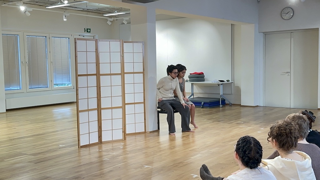 Two actors sit next to each other on a black piano bench in mid-performance of 'Faith Hope Charity'. A small group of students sit watching them to the bottom right of the photo.